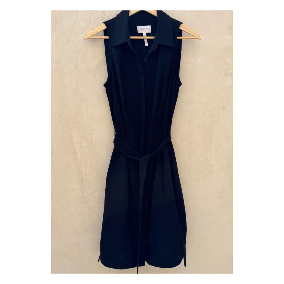 Vestido camisero negro sin mangas Laundry by Shelli Segal con cinturón, modelo de pie descalza sobre fondo cálido tipo chukum, estilo parisino de lujo relajado